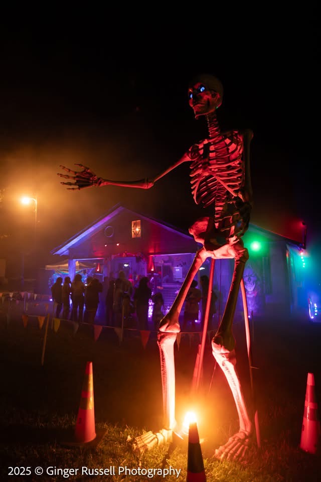 A giant skeleton decoration lit in red and orange towering over the haunted house at night