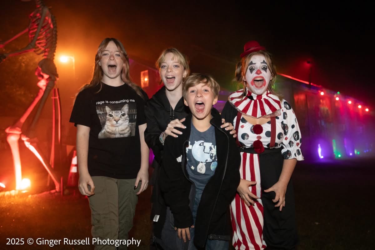 A group of kids grinning and posing with a costumed clown character, string lights in the background