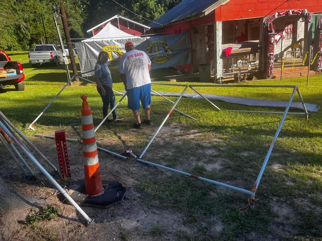 Two Hemphill Lions Club members assembling a tent frame in the yard, with Lions International banners hung on the fence and the haunted house decorated in the background