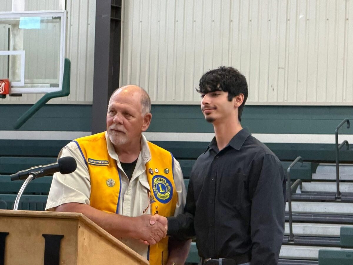 A Hemphill Lions Club member in a gold club vest, Lion of the Year patch, shaking hands with a graduating student at a scholarship presentation