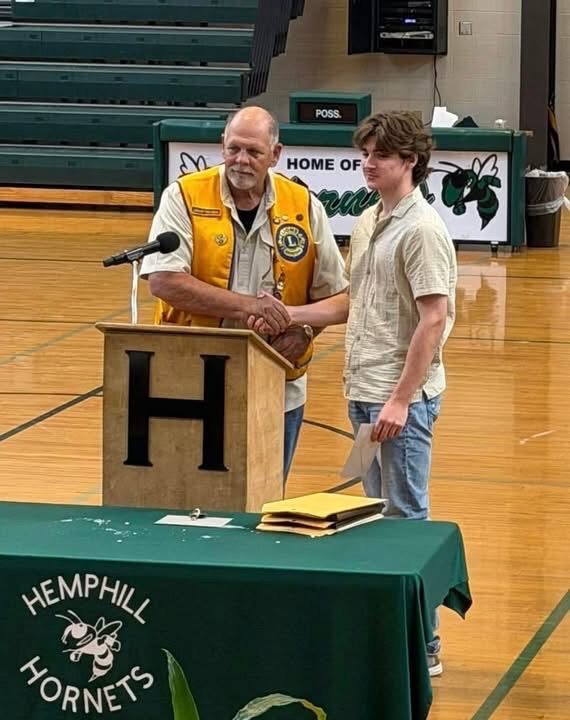 A Hemphill Lions Club member in a gold vest shaking hands with a student at a podium reading H, Hemphill Hornets tablecloth
