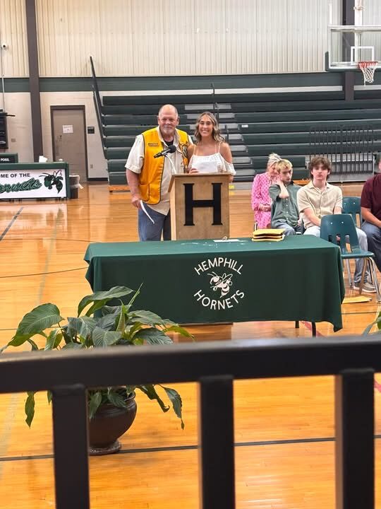 A Hemphill Lions Club member presenting a scholarship to a student at a podium, other recipients seated nearby, Hemphill Hornets gymnasium
