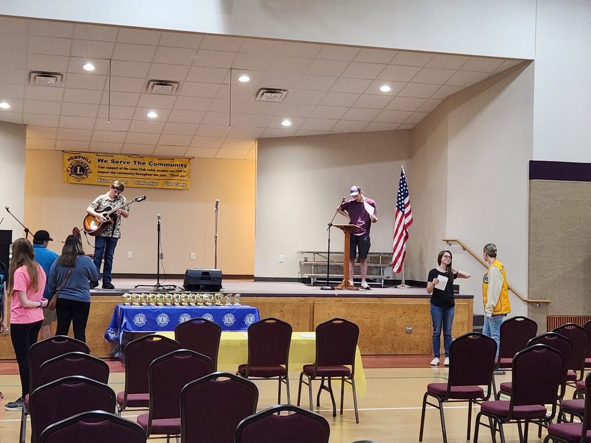 A performer soundchecking on the Talent Show stage during setup, with the club's We Serve The Community banner and a table of trophies at the stage edge